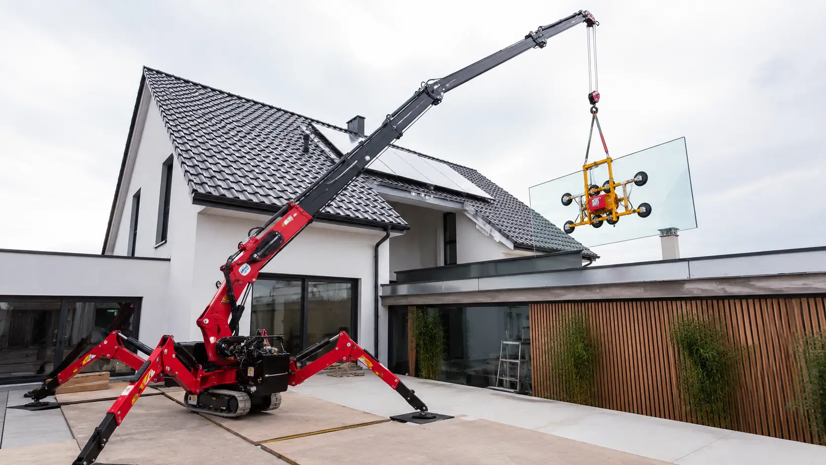 Spinnenkran hebt mit Vakuum-Glassauger eine große Glasscheibe über ein Hausdach in einen Innenhof