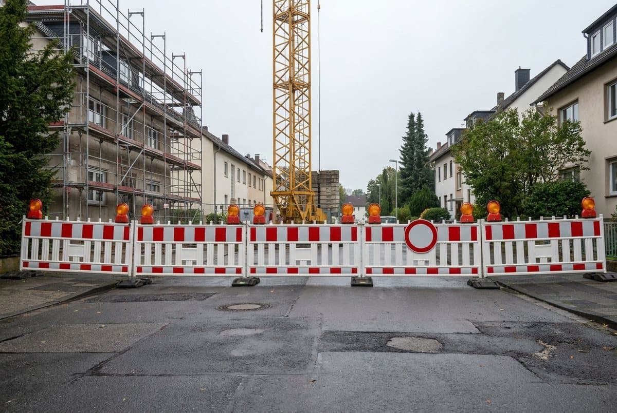 Vollgesperrte Wohnstraße: rot-weiße Absperrschranken mit Warnleuchten quer über die Fahrbahn, dahinter ein eingerüstetes Gebäude und der Gittermast eines gelben Baukrans